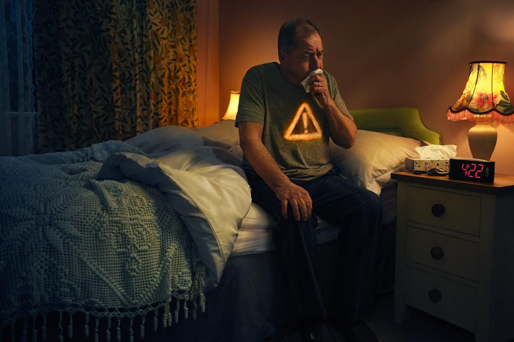 A man in a bed, sitting on the edge, holding a tissue to his nose, with a nightstand and a lamp in the background.