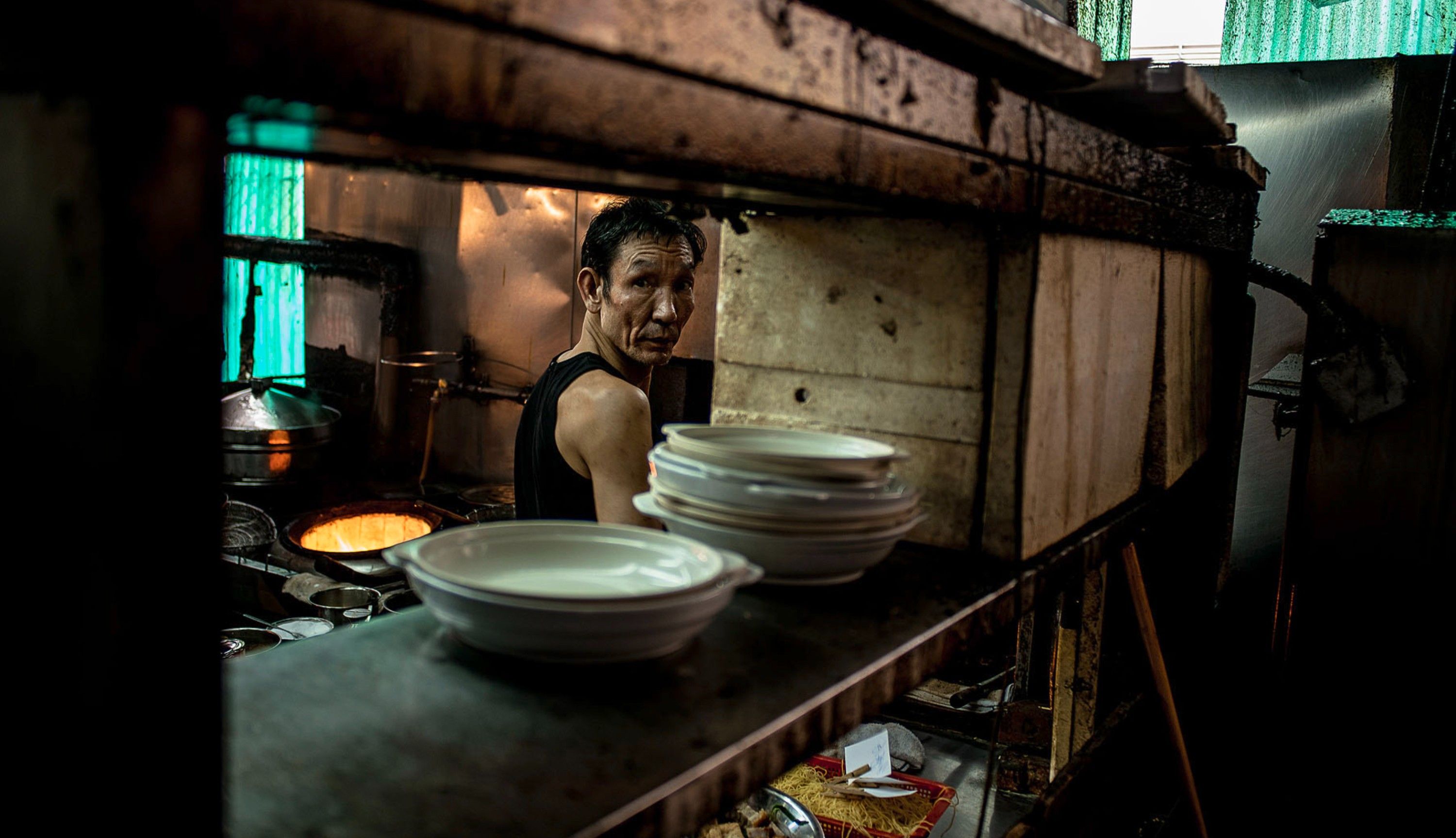 A chef in a black tank top is standing in a dimly lit kitchen, surrounded by cooking equipment and food preparation areas.