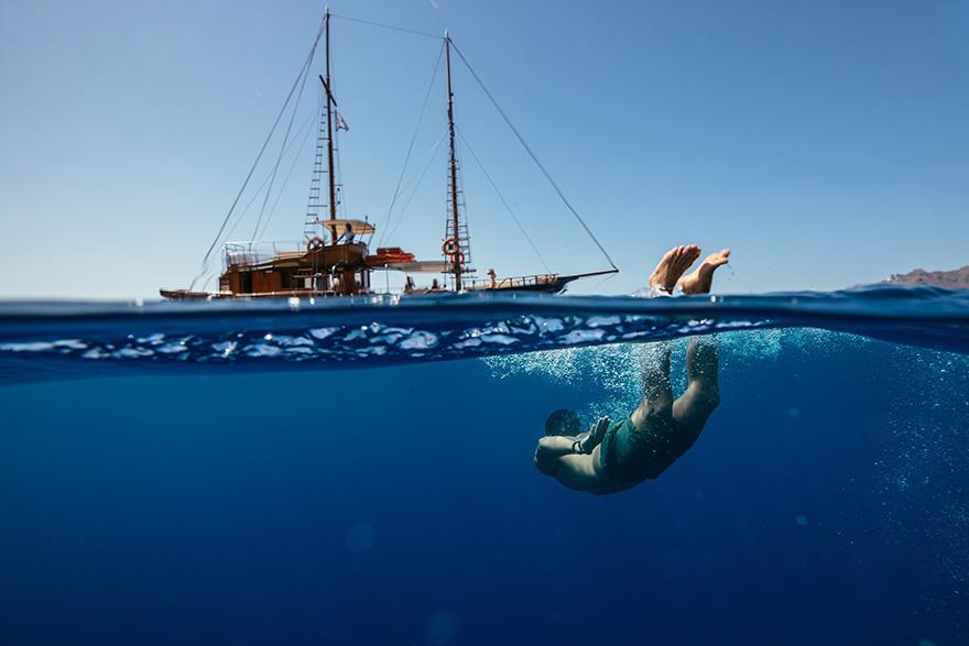 A person in a wetsuit is diving underwater, with a large sailing ship in the background.