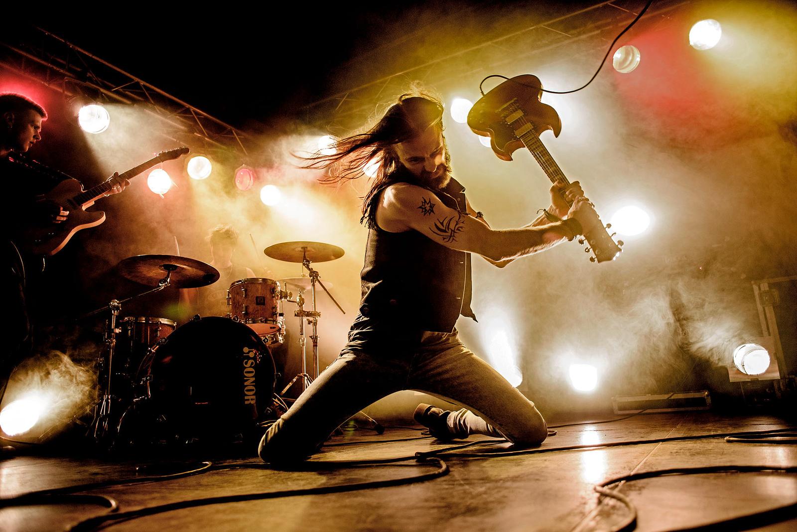 A rock band performs on stage, with a guitarist in the foreground, strumming his instrument with wild hair and a fierce expression. The atmosphere is electric, with a sense of.