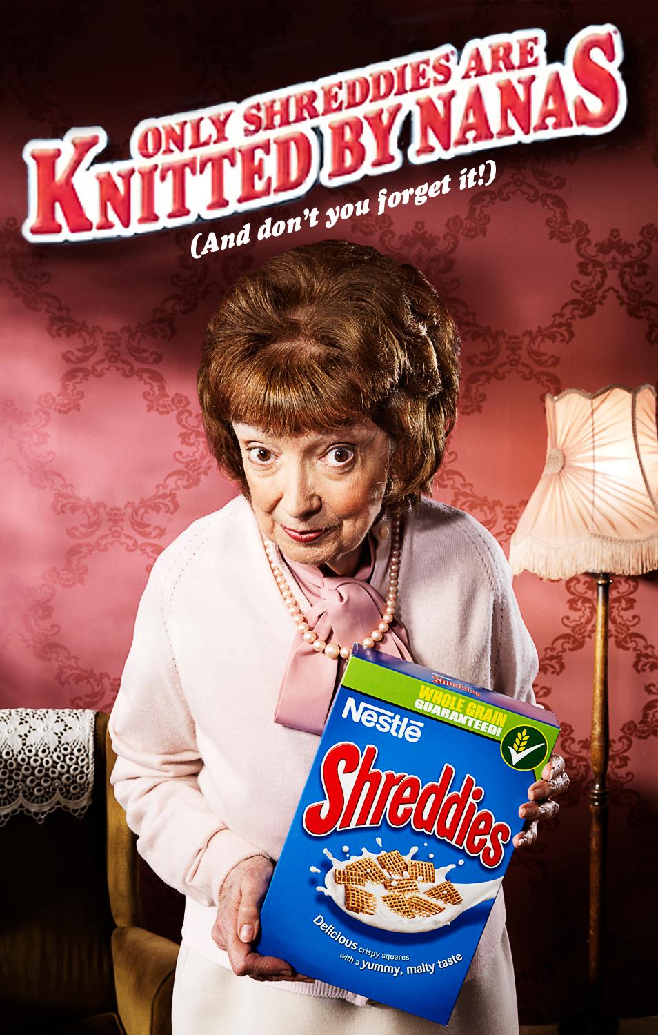 An elderly woman with short brown hair and a white cardigan holds a box of Shreddies cereal. She has a worried and slightly disapproving expression.