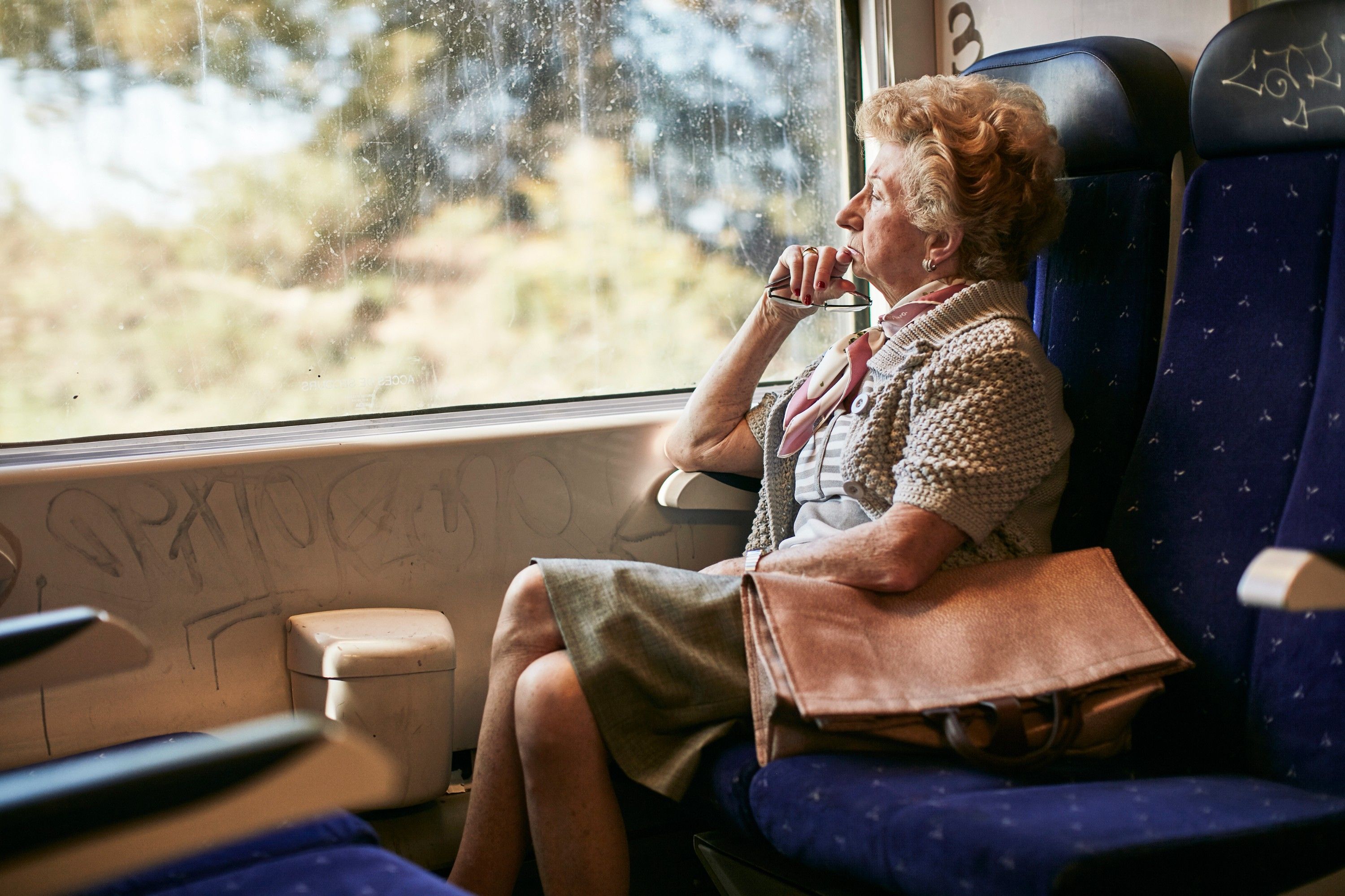An elderly woman sits alone on a train, gazing out the window at a blurred landscape.