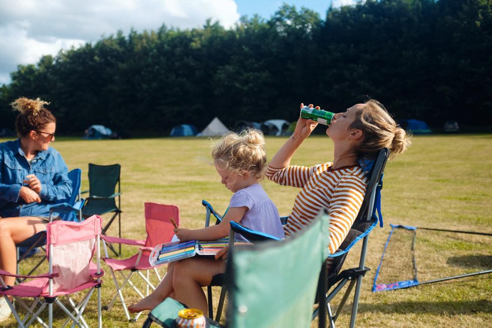 A woman in a striped shirt sits in a chair, holding a green can and drinking from it, while a young girl sits beside her, coloring in a book. In the background, a grassy field.