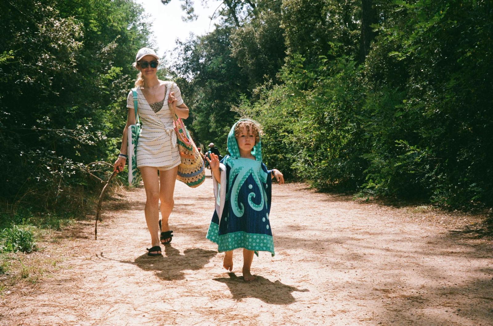 A woman and a young child walk down a dirt path in a forest, both wearing casual summer outfits. The woman is carrying a large bag and a stick, while the child is covered in a.
