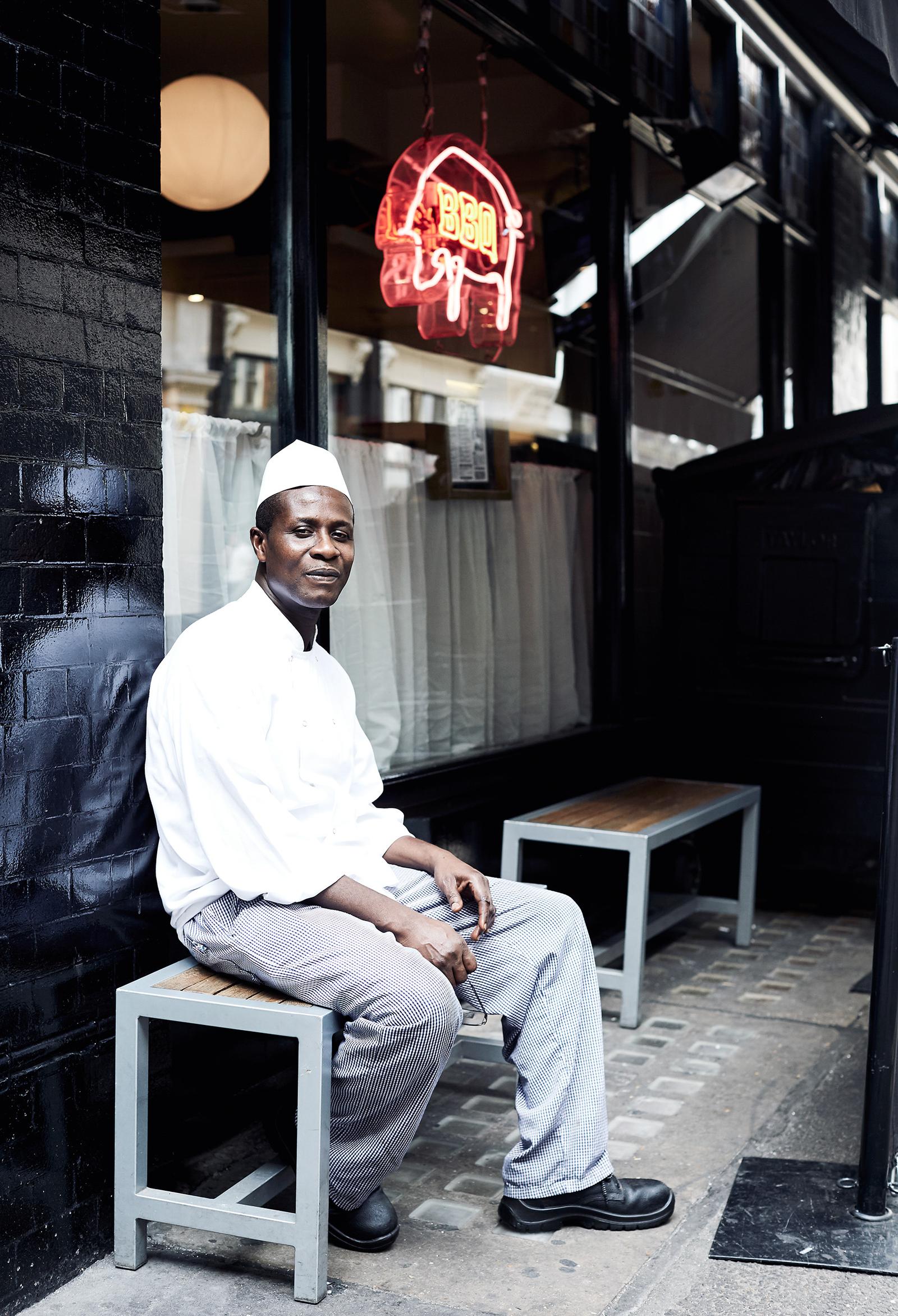 A man in a white chef's hat and white shirt sitting on a stool outside a restaurant, likely in a city or urban setting.