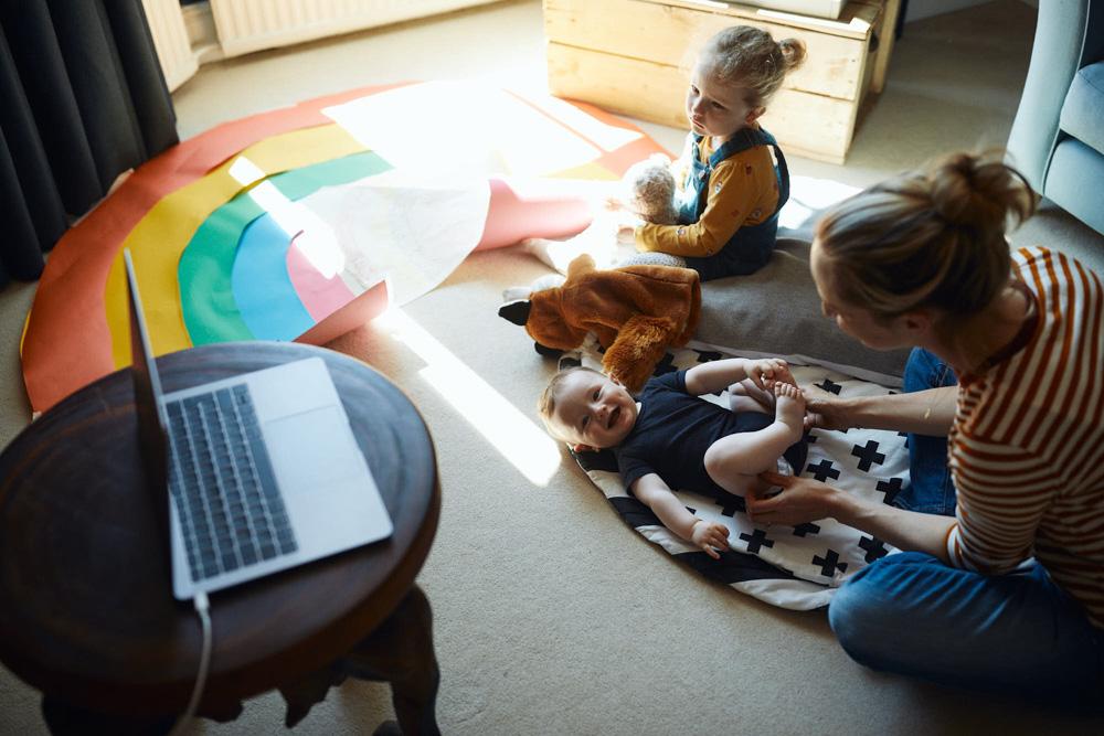 A young woman with a child and an infant on the floor, surrounded by toys and a laptop. The woman is smiling, while the child is playing with a toy, and the infant is lying on a.