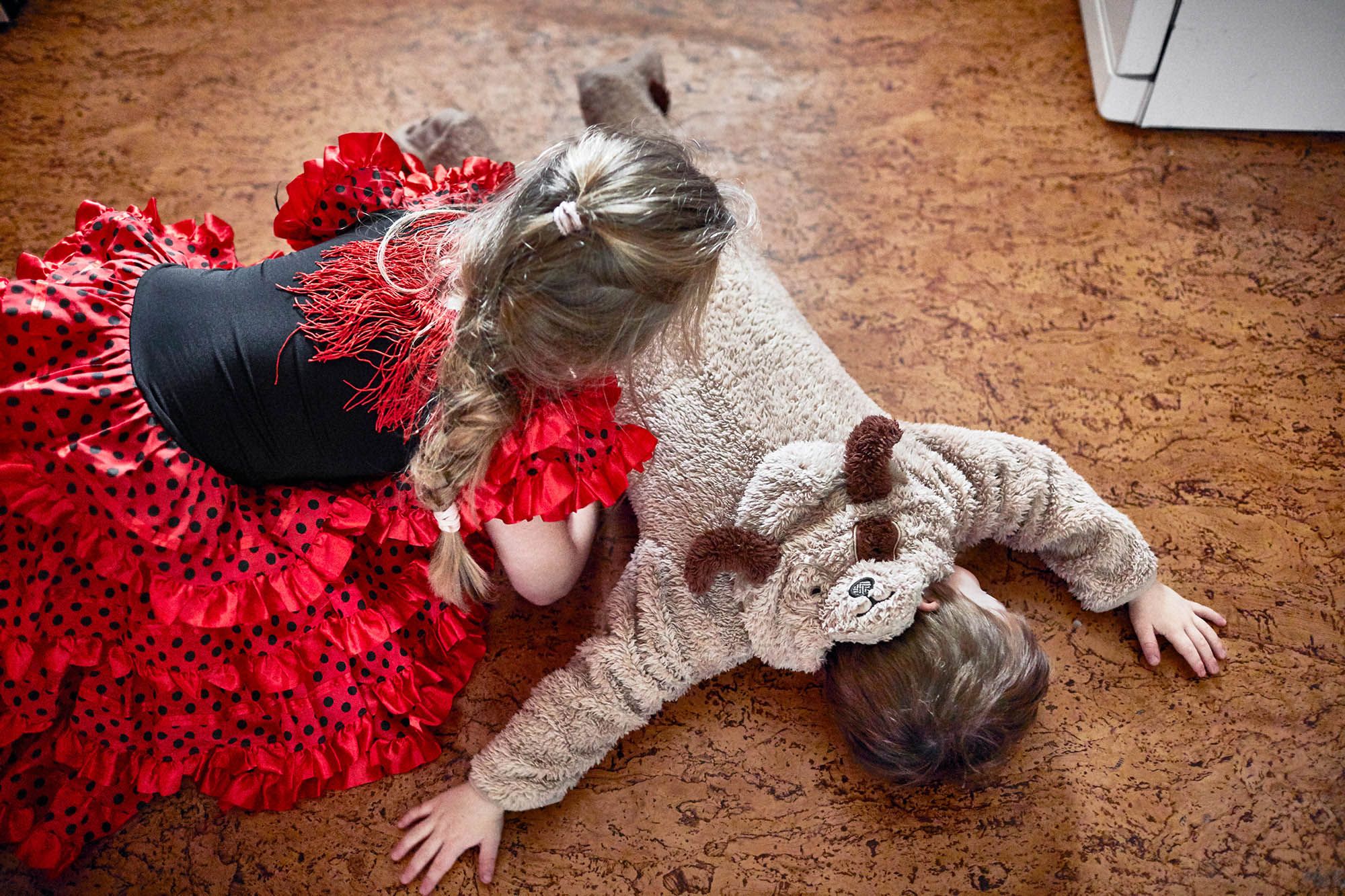 A young girl in a red flamenco dress and a boy in a teddy bear onesie are playing on the floor.