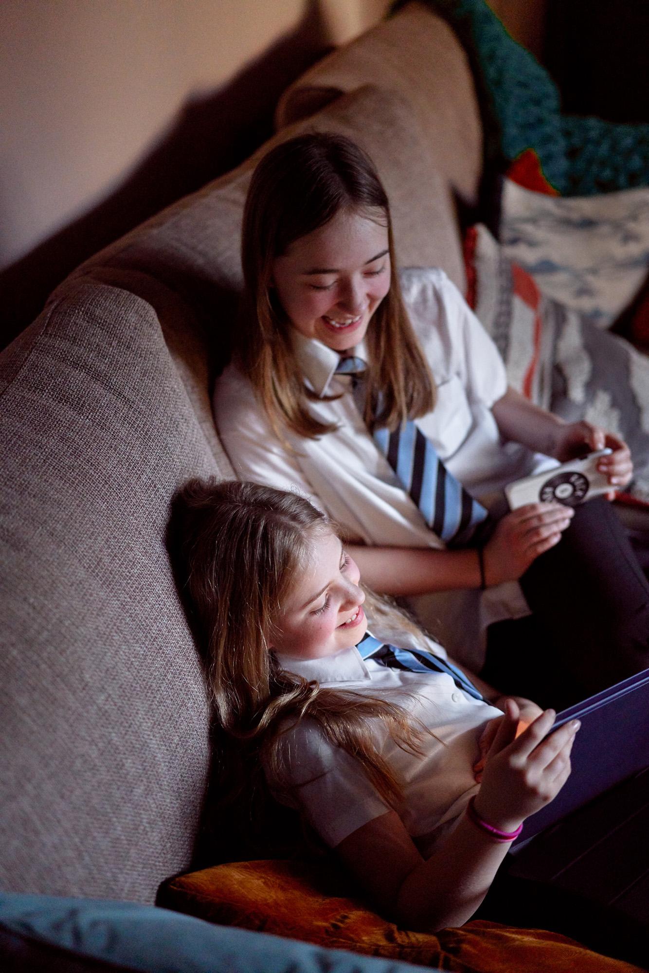 Two girls in school uniforms sitting on a couch, one holding a book and the other holding a device.