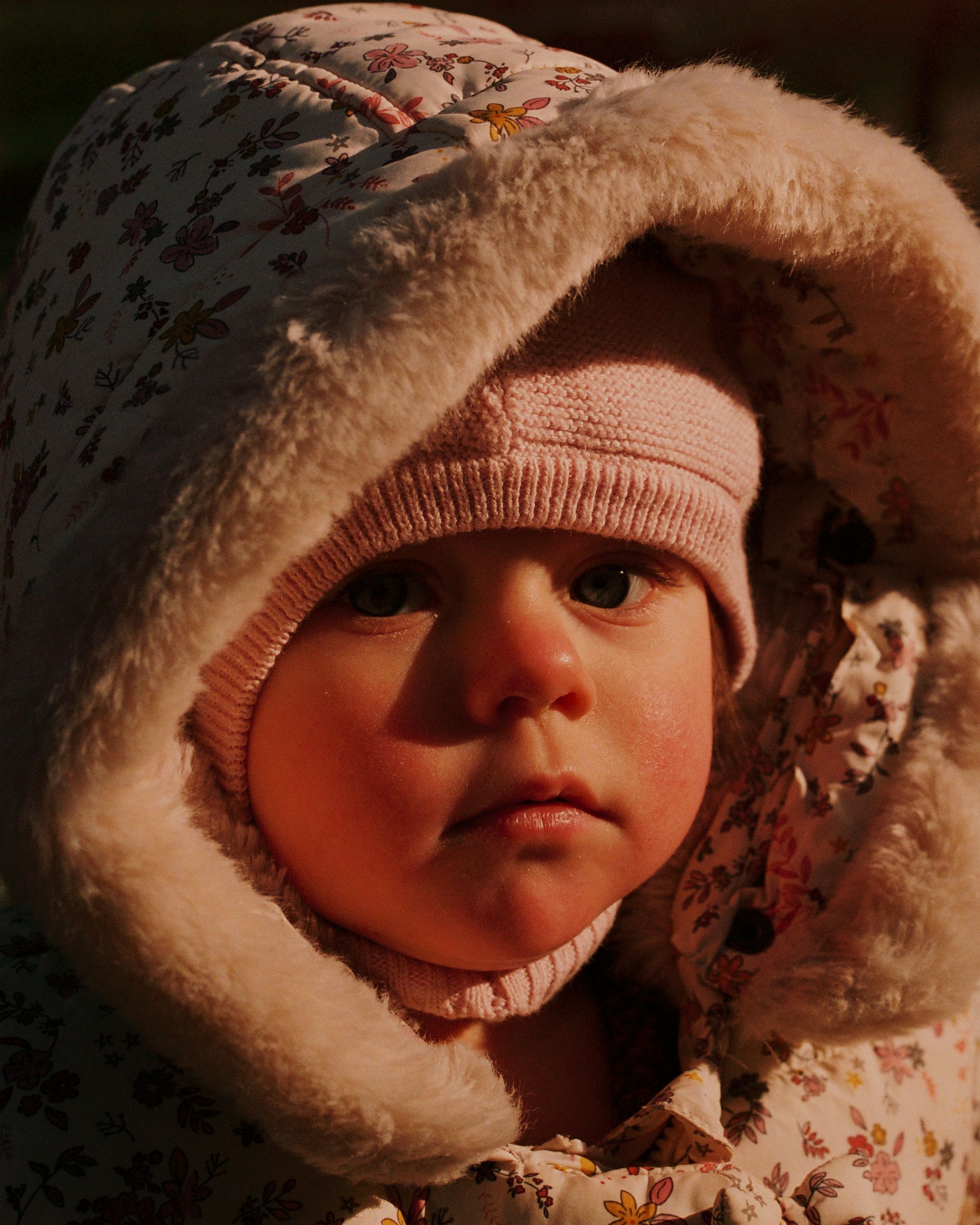 A baby in a pink hat and coat, with a fur-lined hood, looking directly at the camera with a serious expression.