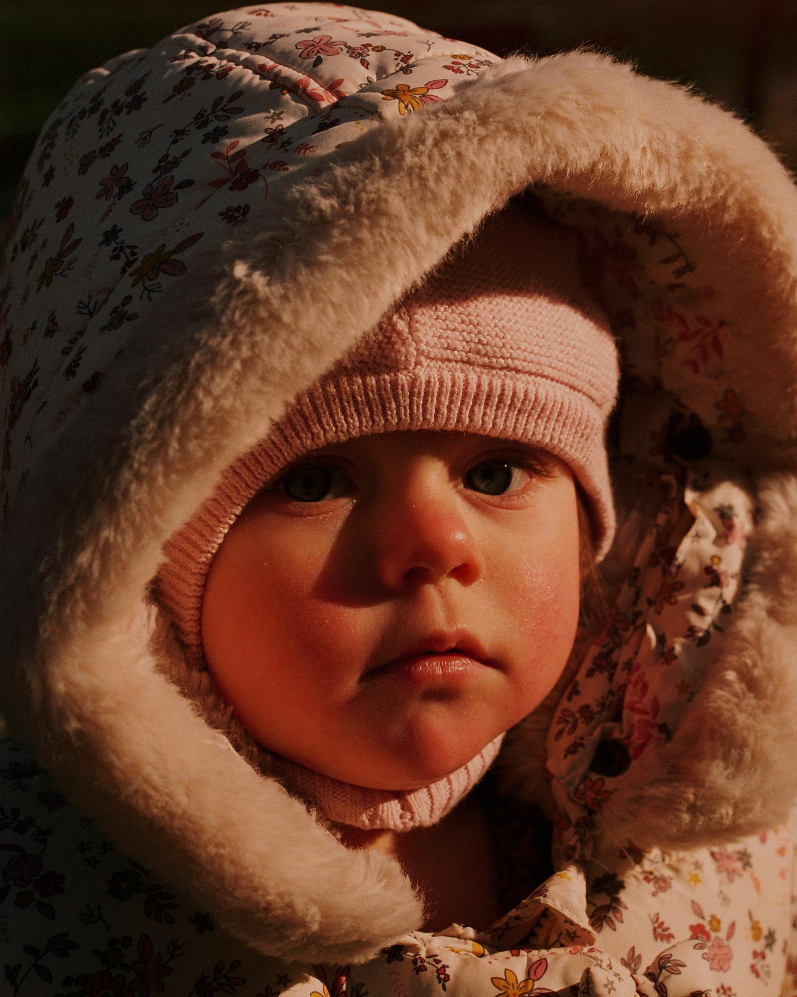 A baby in a pink hat and coat, with a fur-lined hood, looking directly at the camera with a serious expression.