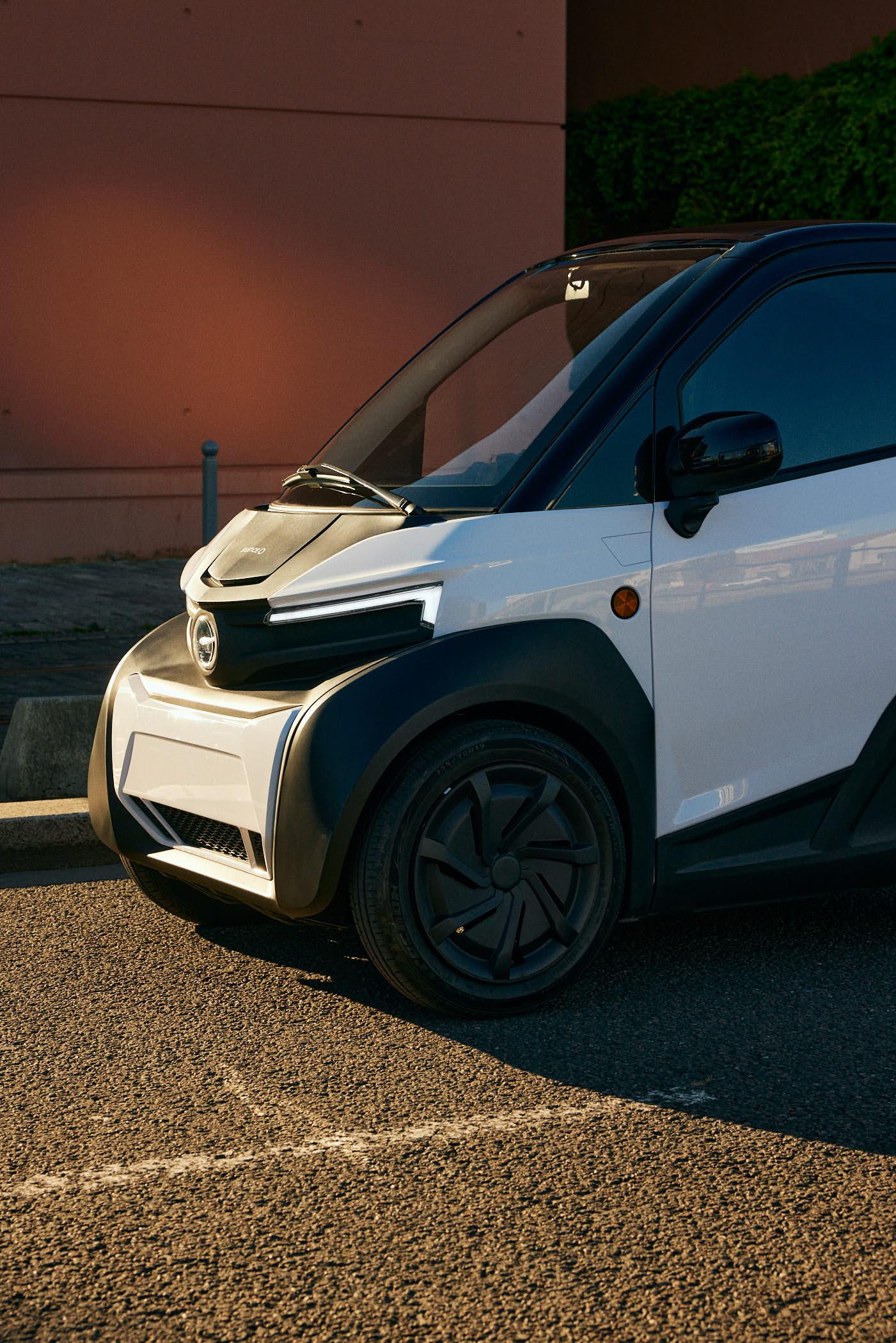 A small, white, electric car is parked in front of a red wall, likely in a parking lot or parking space. The car's design is modern and compact, with a curved, aerodynamic shape.