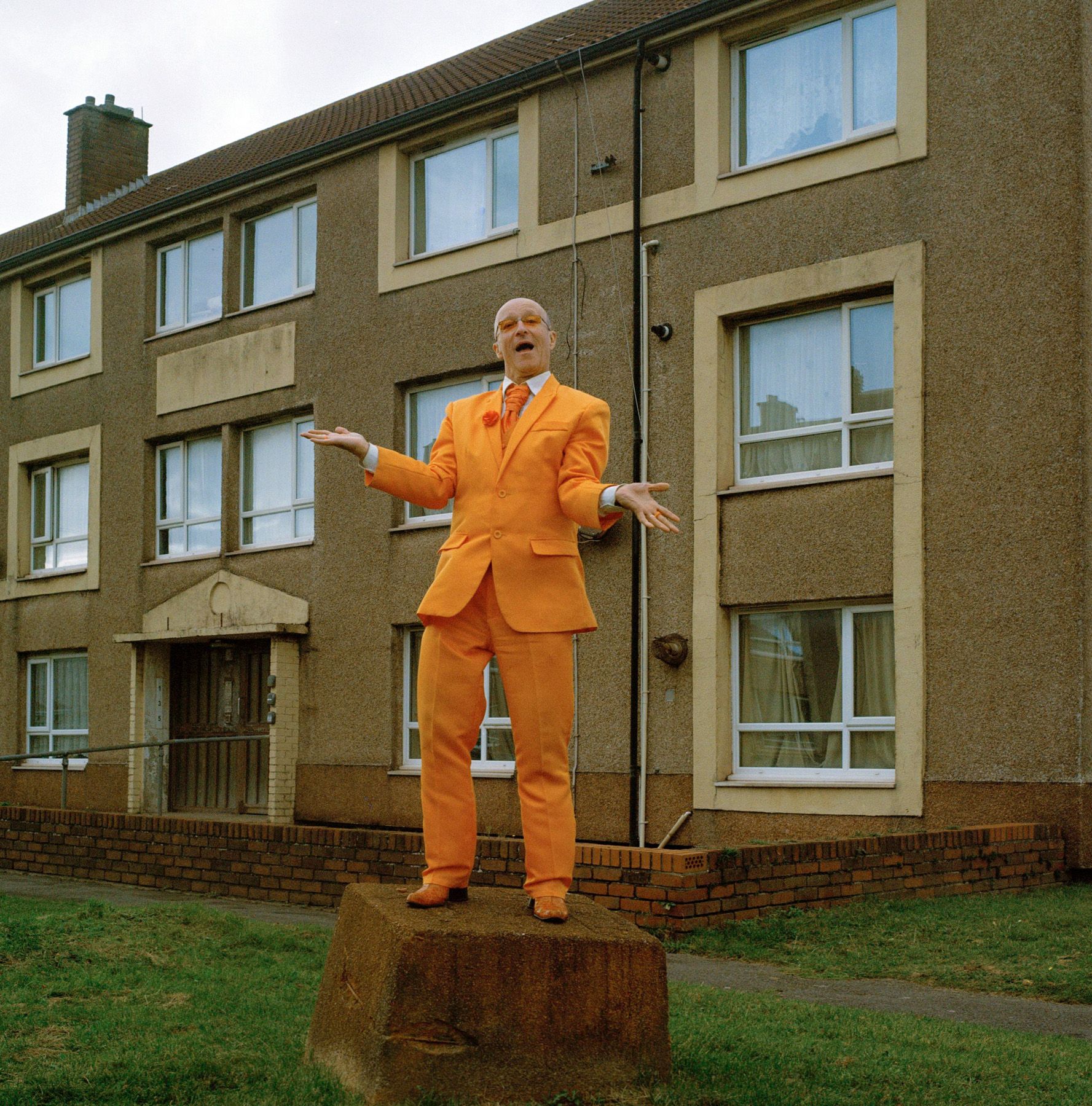 A man in an orange suit stands on a large stone in front of old brick buildings.