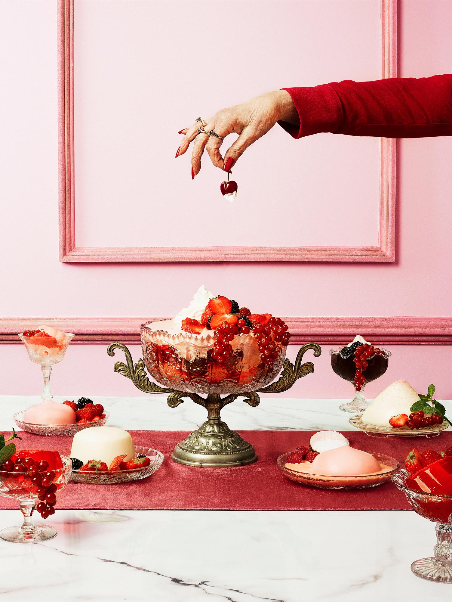 A hand reaches for a cherry among sweet treats on a dessert table.