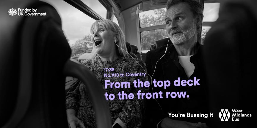 A woman with a big smile on her face, wearing a patterned dress, sitting in a bus, with a man in the background, looking at her.