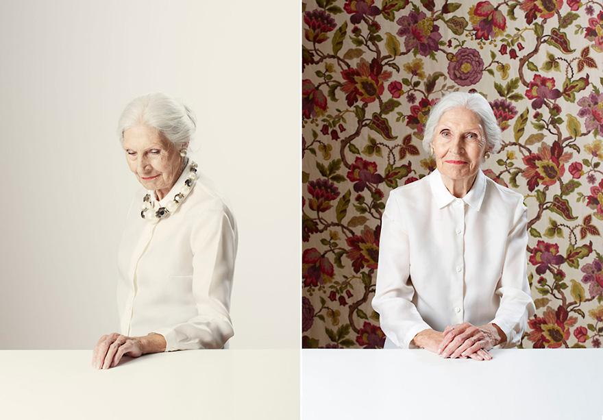 An elderly woman with white hair, wearing a white blouse with a high collar and a patterned scarf, standing in front of a floral wallpaper.
