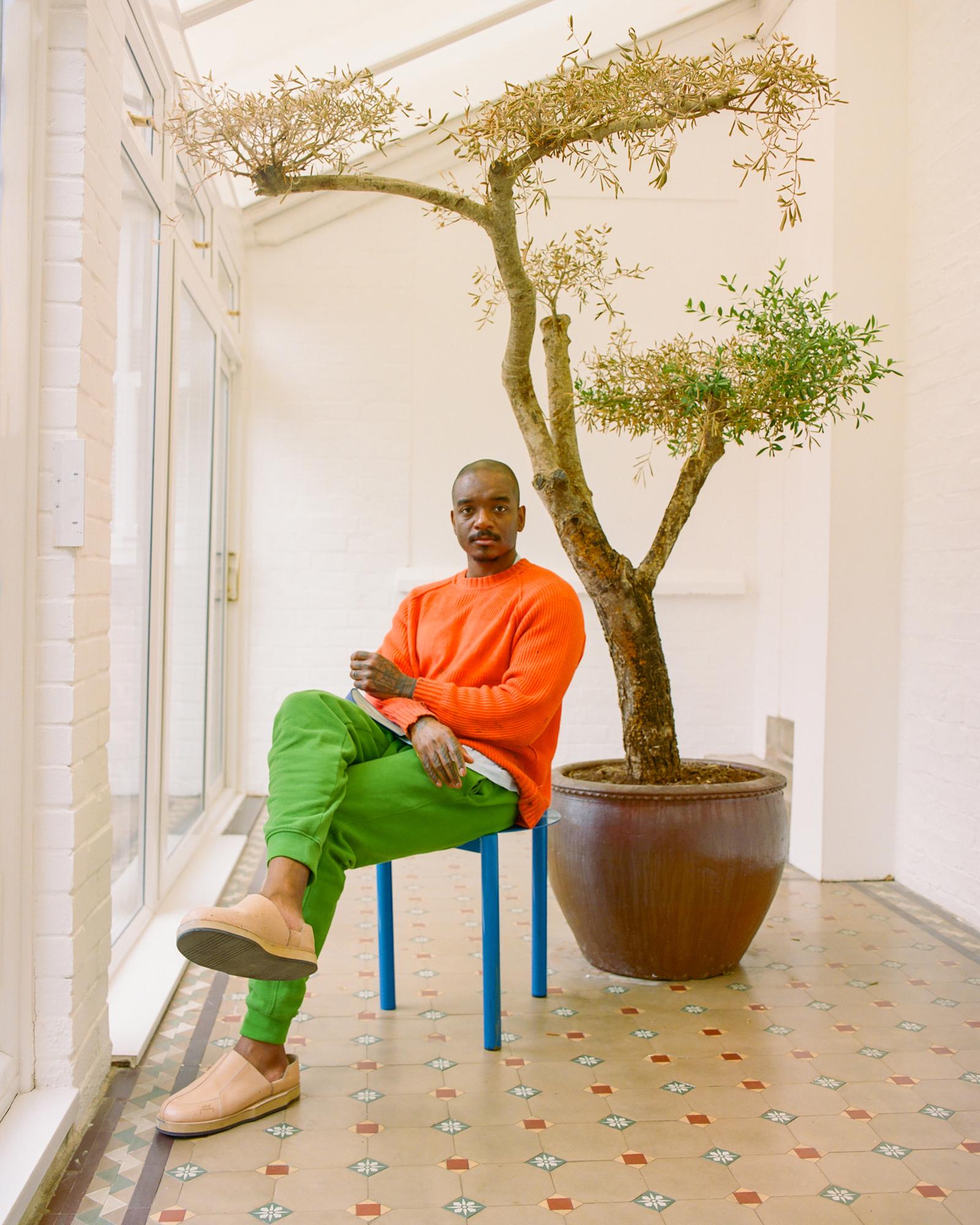 A man sitting on a chair in a room with a large window, wearing an orange sweater, green pants, and brown shoes.