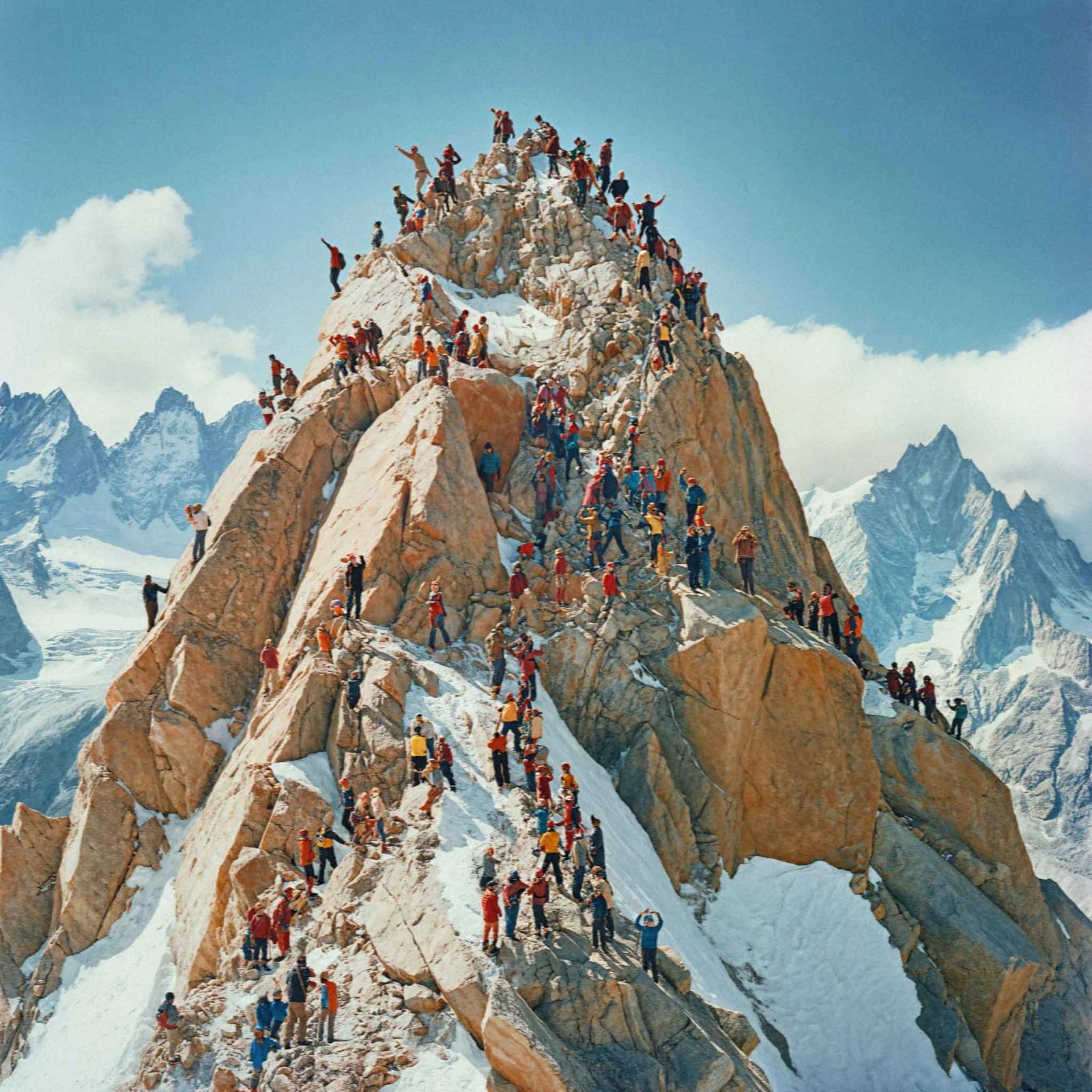 A large crowd of people standing on top of a mountain peak, with some people waving and others taking photos.