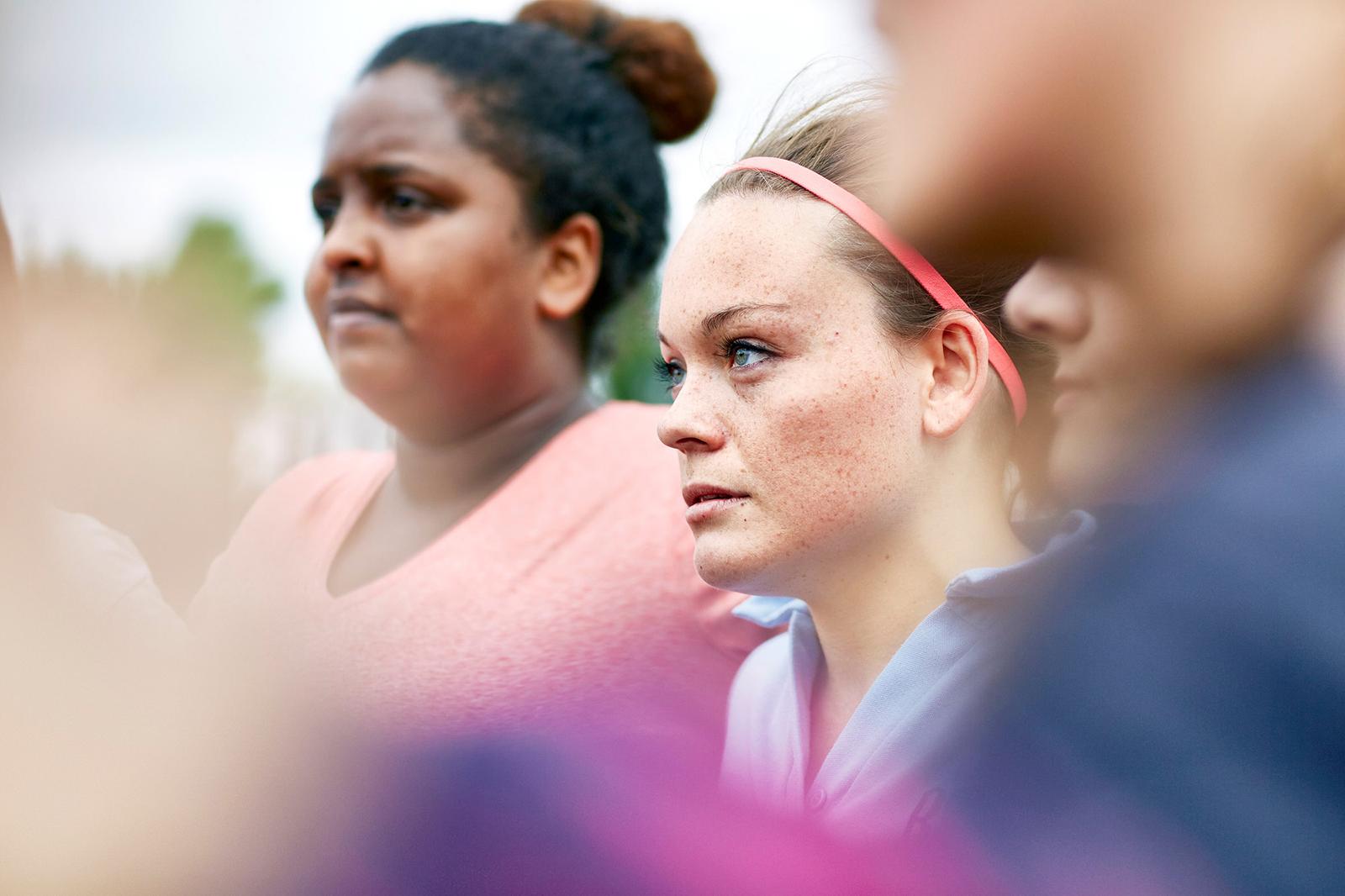 A group of diverse young women, including one with a visible disability, standing together in a park or outdoor setting, wearing casual clothing such as t-shirts and jeans.