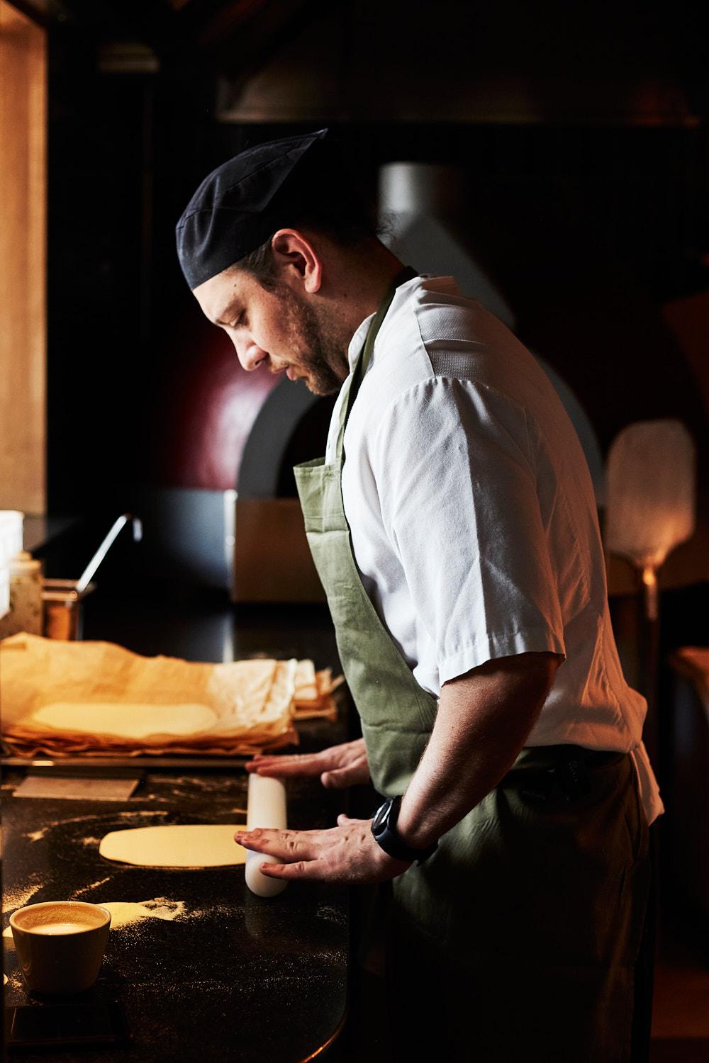 A chef in a white shirt and green apron is expertly rolling out a large piece of pasta dough on a black marble counter, with a few flour-covered hands in the background.