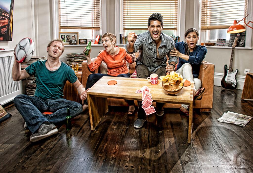 A group of people sitting on a couch, laughing and cheering with a football in the air, surrounded by snacks and drinks.