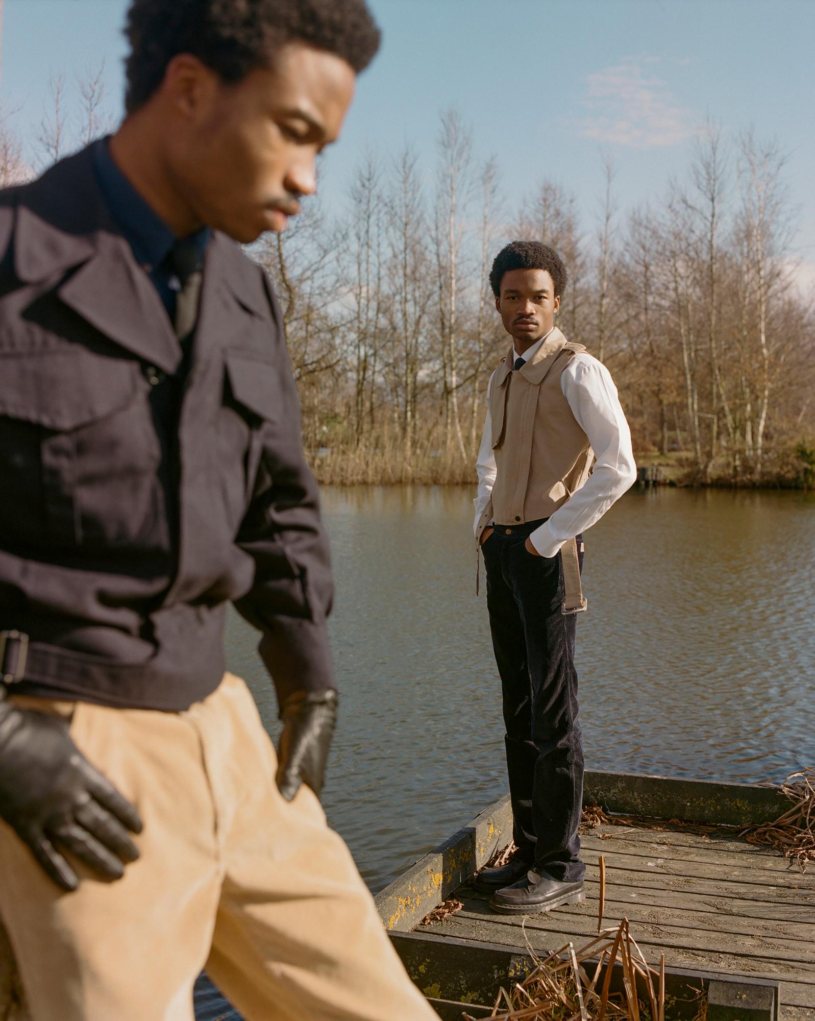 Two men in formal attire standing on a dock overlooking a body of water, with one man in the foreground and the other in the background.