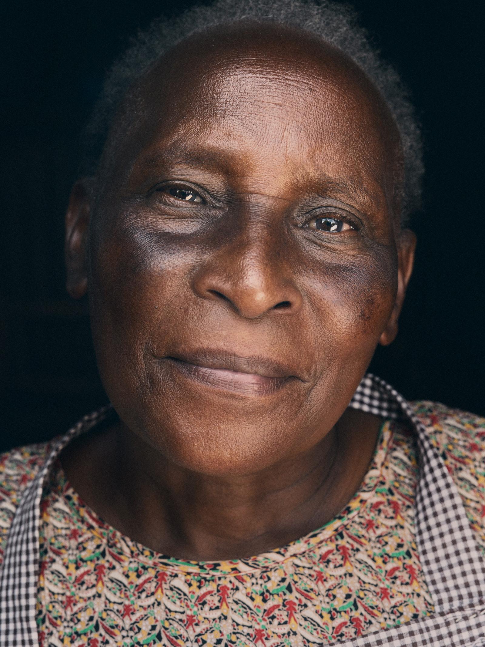 A close-up portrait of an elderly woman with a serious expression, wearing a patterned red, green, yellow, and white shirt with a black and white checkered collar.