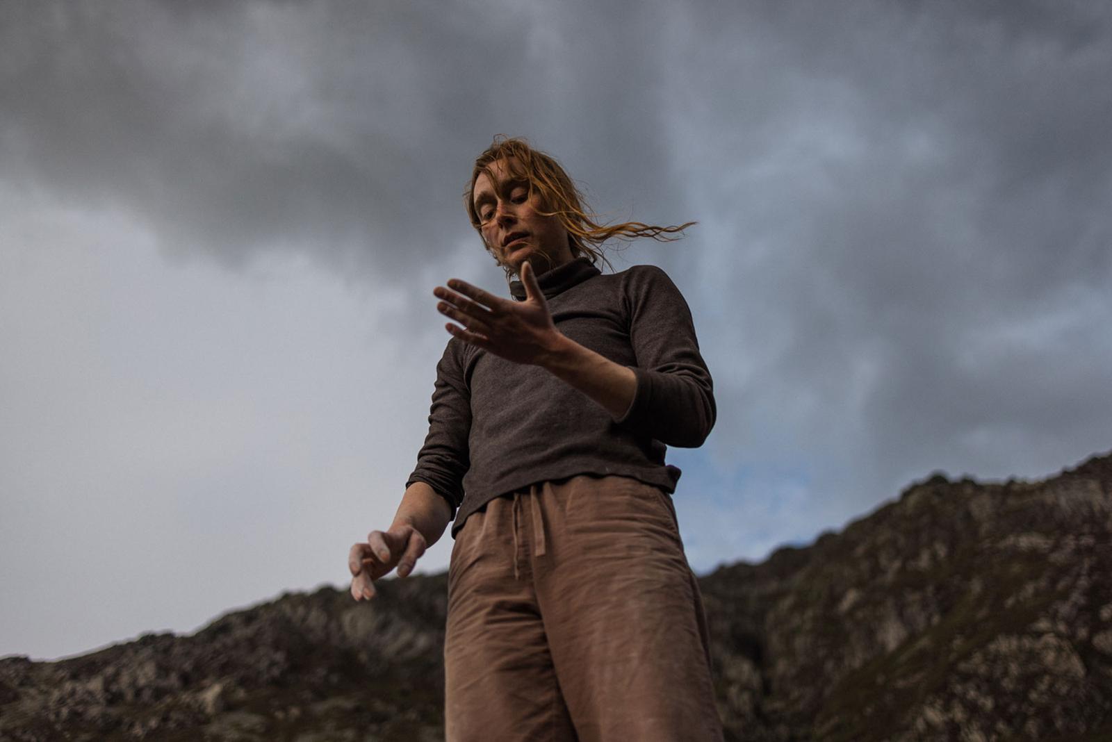 A man with long, wind-blown hair and a grey turtleneck sweater stands in front of a rugged, rocky landscape, with his arms outstretched and his face looking up towards the sky.