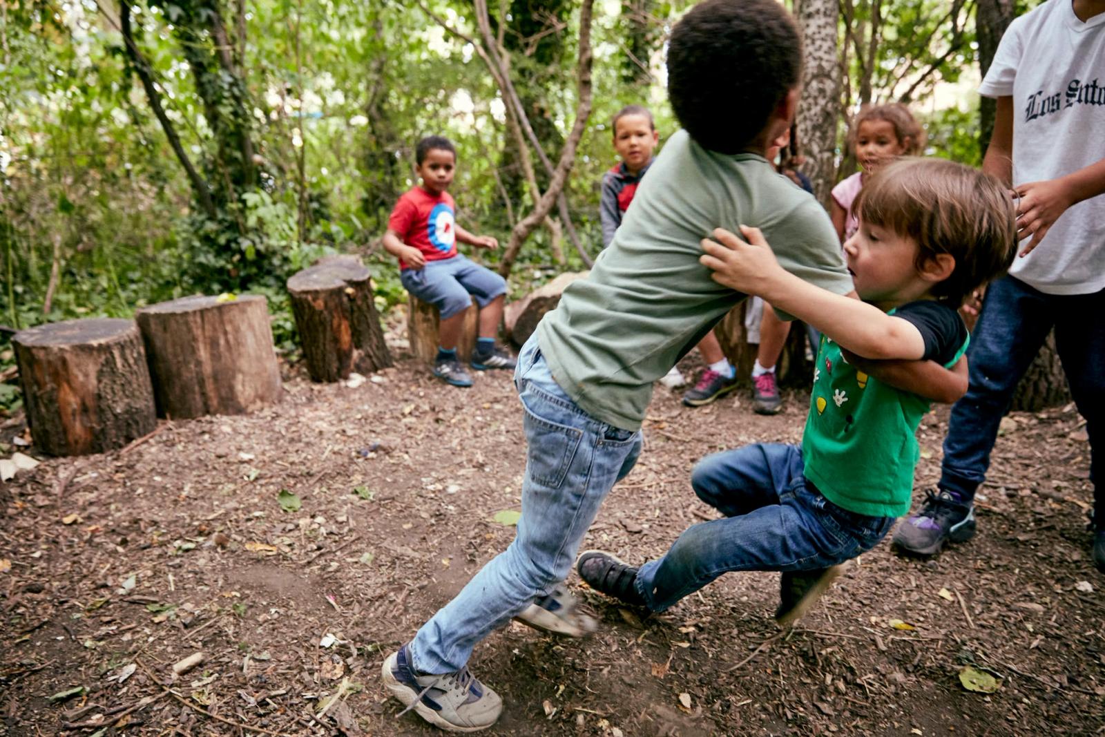 A group of children playing in a forested area, with one child in the foreground being held by another. The children are all wearing casual clothing and appear to be having fun..