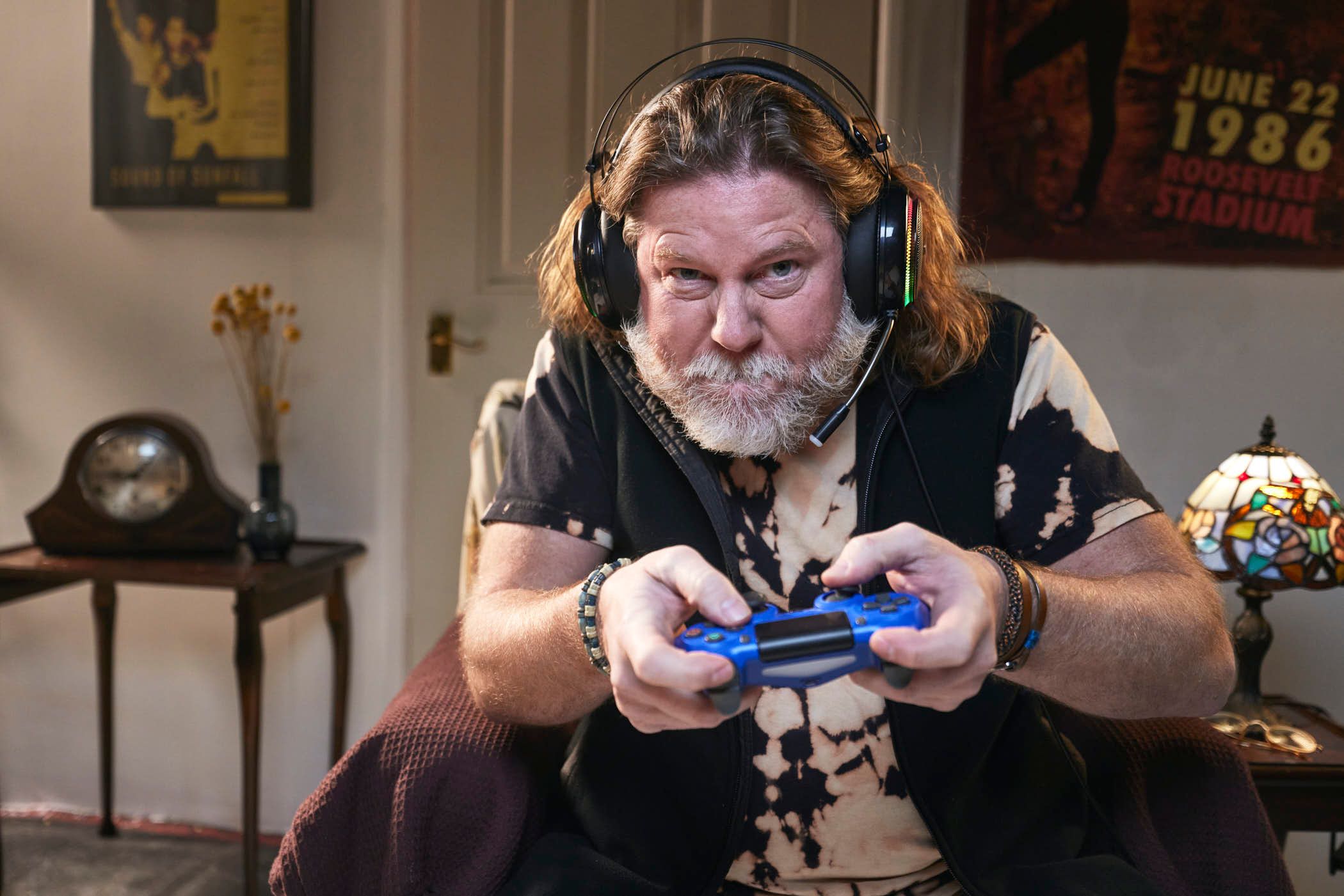 A man with a beard and long hair is sitting in a chair, wearing a headset and holding a video game controller. He is looking at the camera with a serious expression.