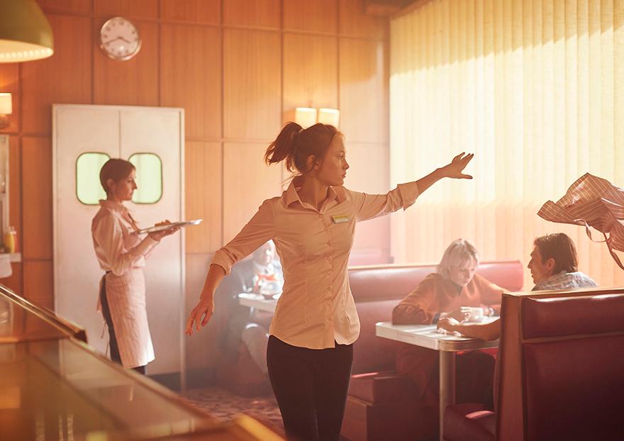 A woman stretches her arms in a diner with a clock on the wall and other patrons nearby.