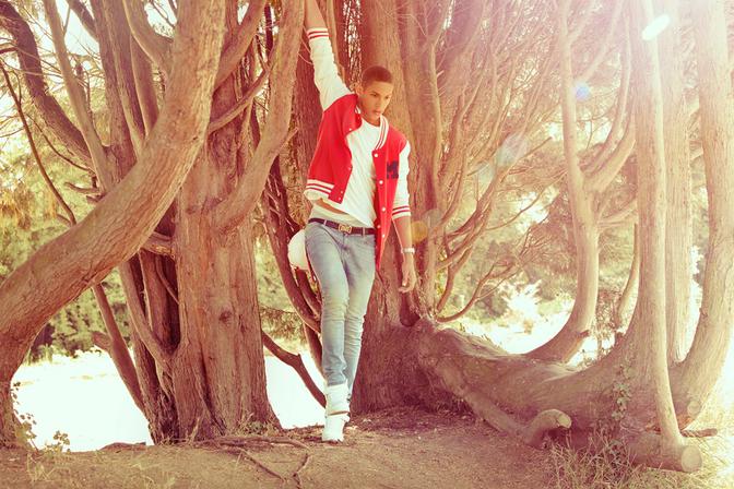 A young man in a red and white striped rugby jersey, jeans, and white sneakers is standing in front of a large tree, with his right arm raised and his left hand in his pocket.