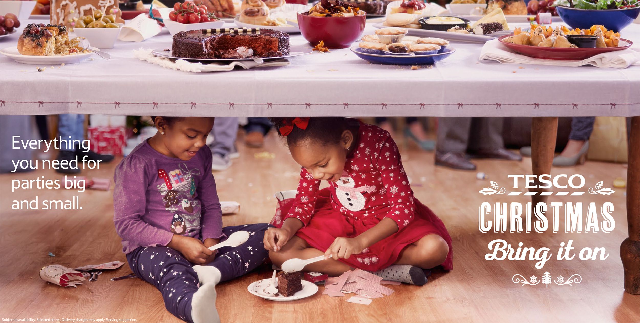 Two young girls sitting on the floor under a table, surrounded by food and food-related items, eating a chocolate cake.