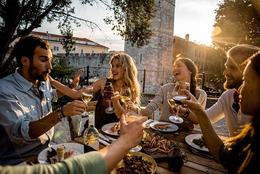 Tourists toasting with wine and enjoying a meal outdoors near historical buildings in Croatia.