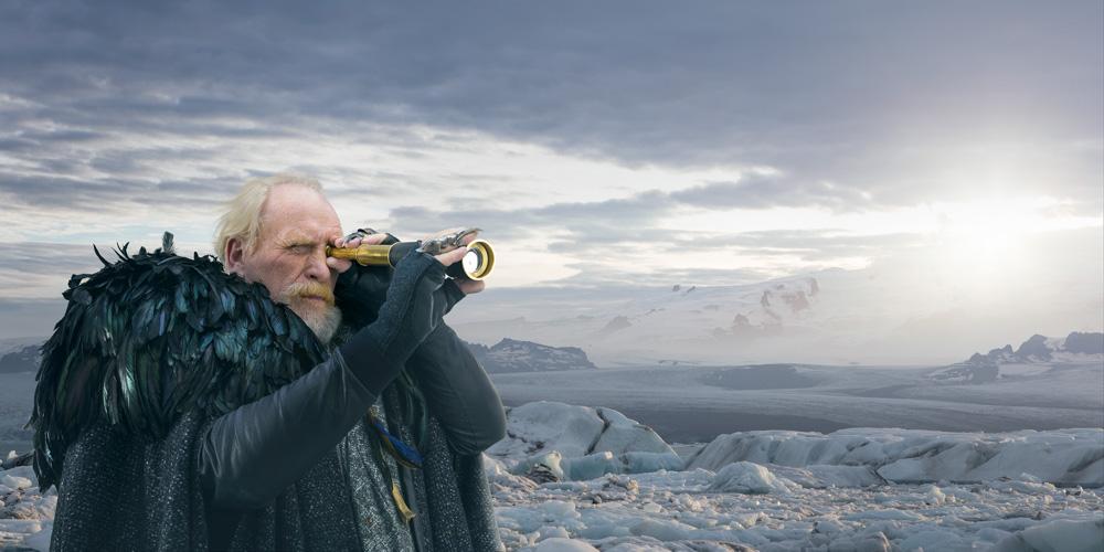 A man with a long white beard and a black feathered cloak, holding a spyglass and standing on a rocky, snow-covered landscape.