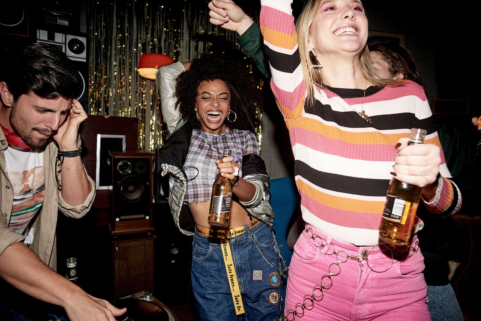 A group of people dancing and celebrating in a dimly lit room, with one person holding a beer bottle.