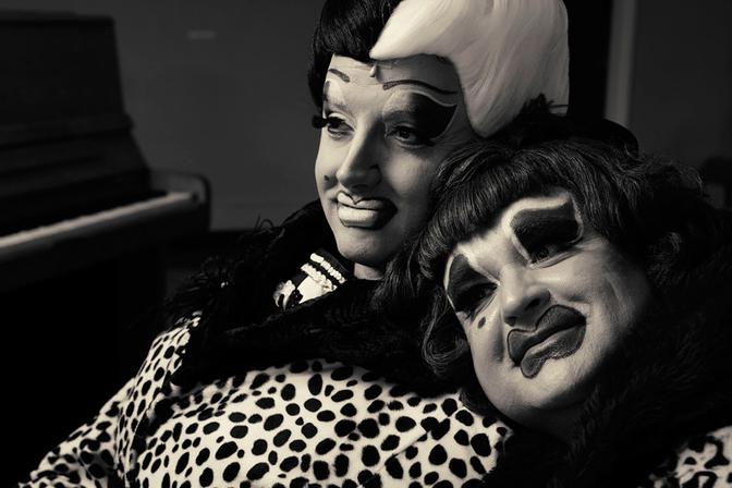 Two drag performers, The Ugly Sisters, dressed in elaborate costumes, including a leopard-print jacket and a pink wig, posing together with a playful and confident expression.