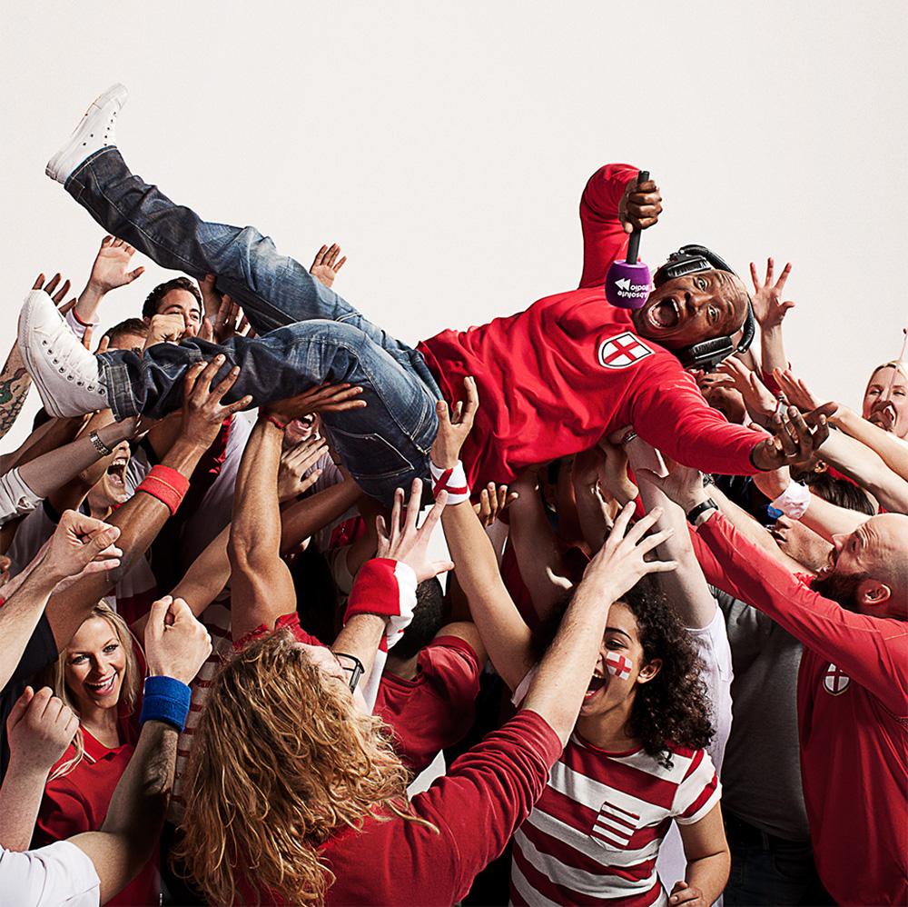 A crowd of people lifting a man in the air, with many hands and arms visible, and the man is wearing a red shirt with a white cross on the chest, and jeans.