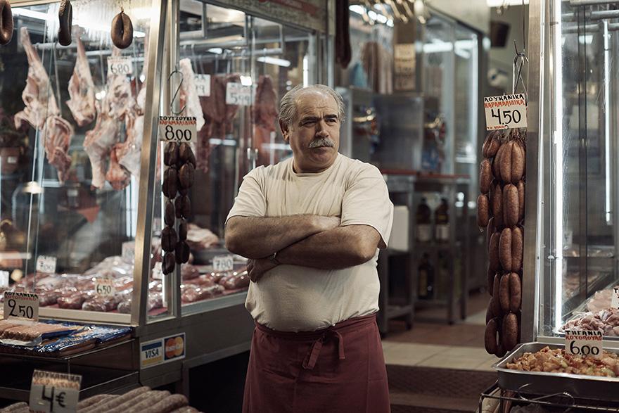 A man with a mustache and a white shirt standing in front of a meat market, surrounded by various types of meat and meat products.
