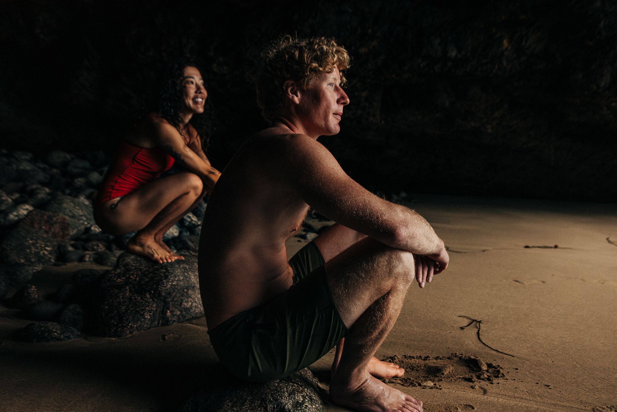 A man and a woman in swimsuits sitting on a rock at the beach, looking out at the ocean.