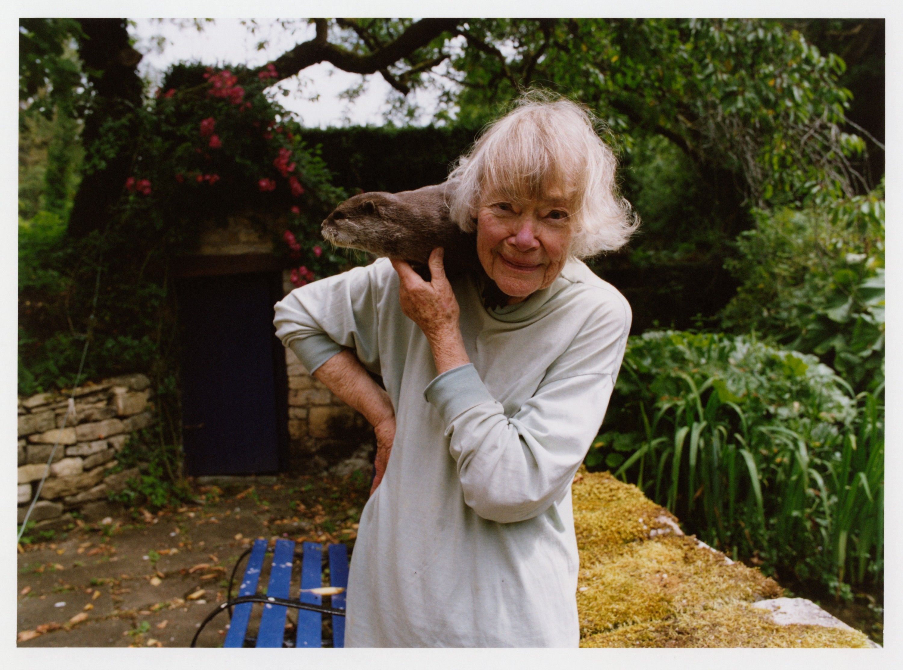 An older woman with short, white hair and a white, long-sleeved turtleneck is holding an otter on her shoulder, with her hand on the otter's back, in a garden setting.