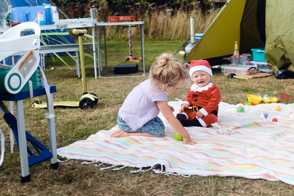 A young girl and a baby in a Santa suit play on a blanket in a campsite. The girl is helping the baby with a green ball, both smiling.