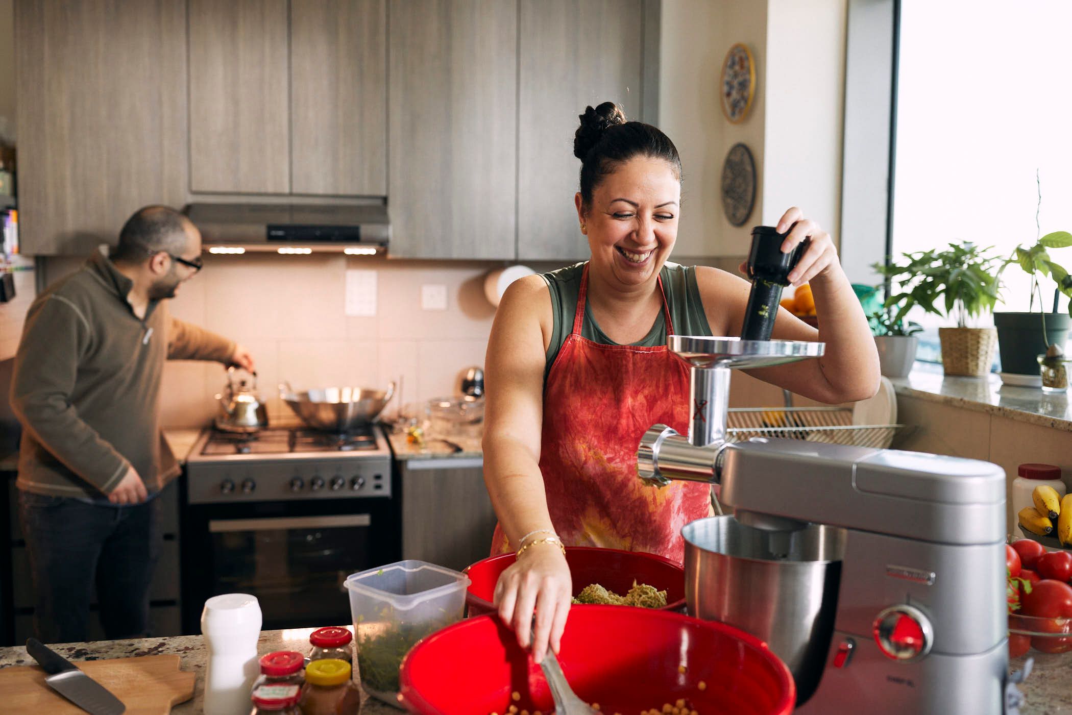 A woman in a red apron is smiling while using a food grinder in a well-lit kitchen. A man is standing at a nearby stove, stirring a pot.