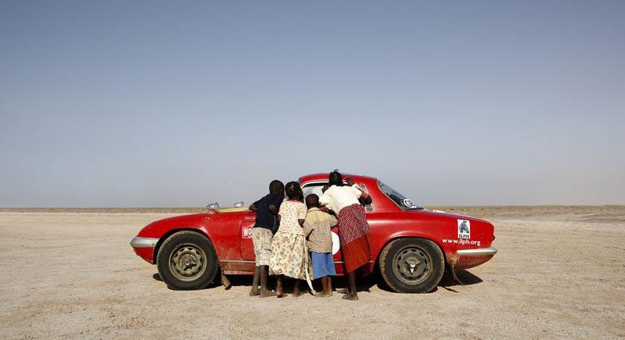 Children gather around a red car in a desert setting.
