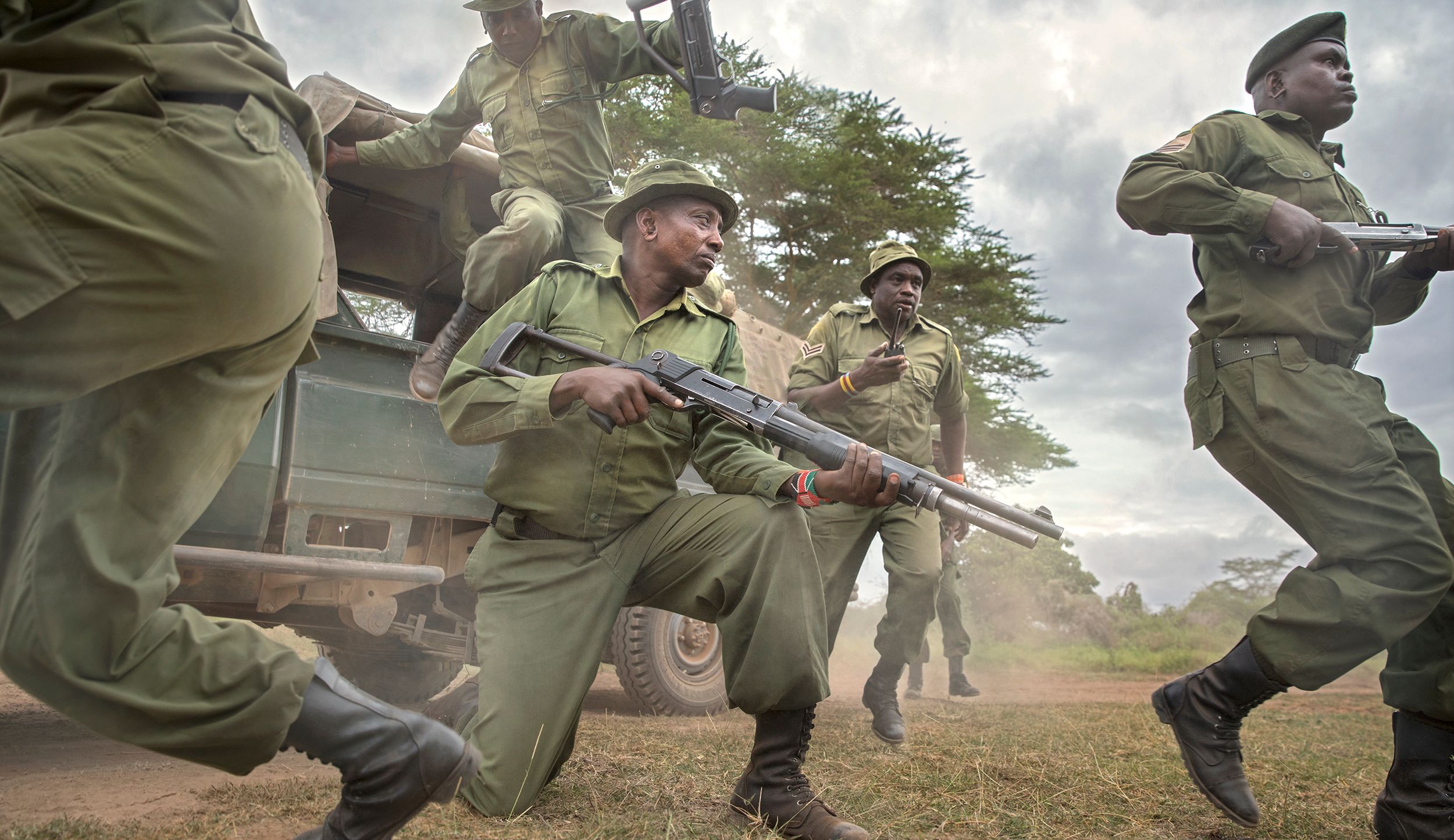 A group of Kenyan wildlife rangers in military-style uniforms, armed with rifles, are on patrol in a forested area, likely in a national park or wildlife reserve.