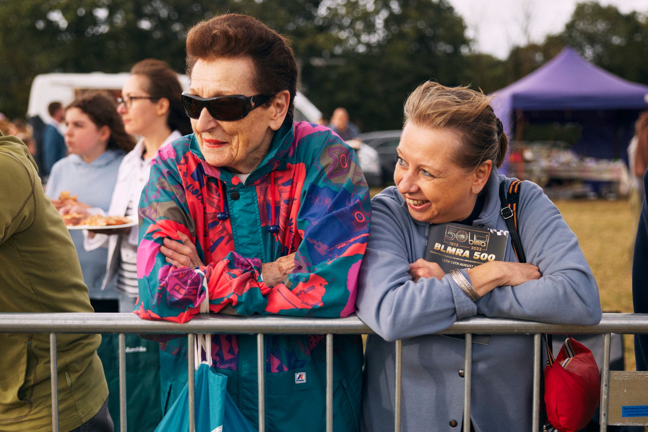 Two elderly women, both with short, styled hair, lean on a metal railing, gazing at something out of frame. The woman on the left wears a vibrant, multicolored jacket, while the.