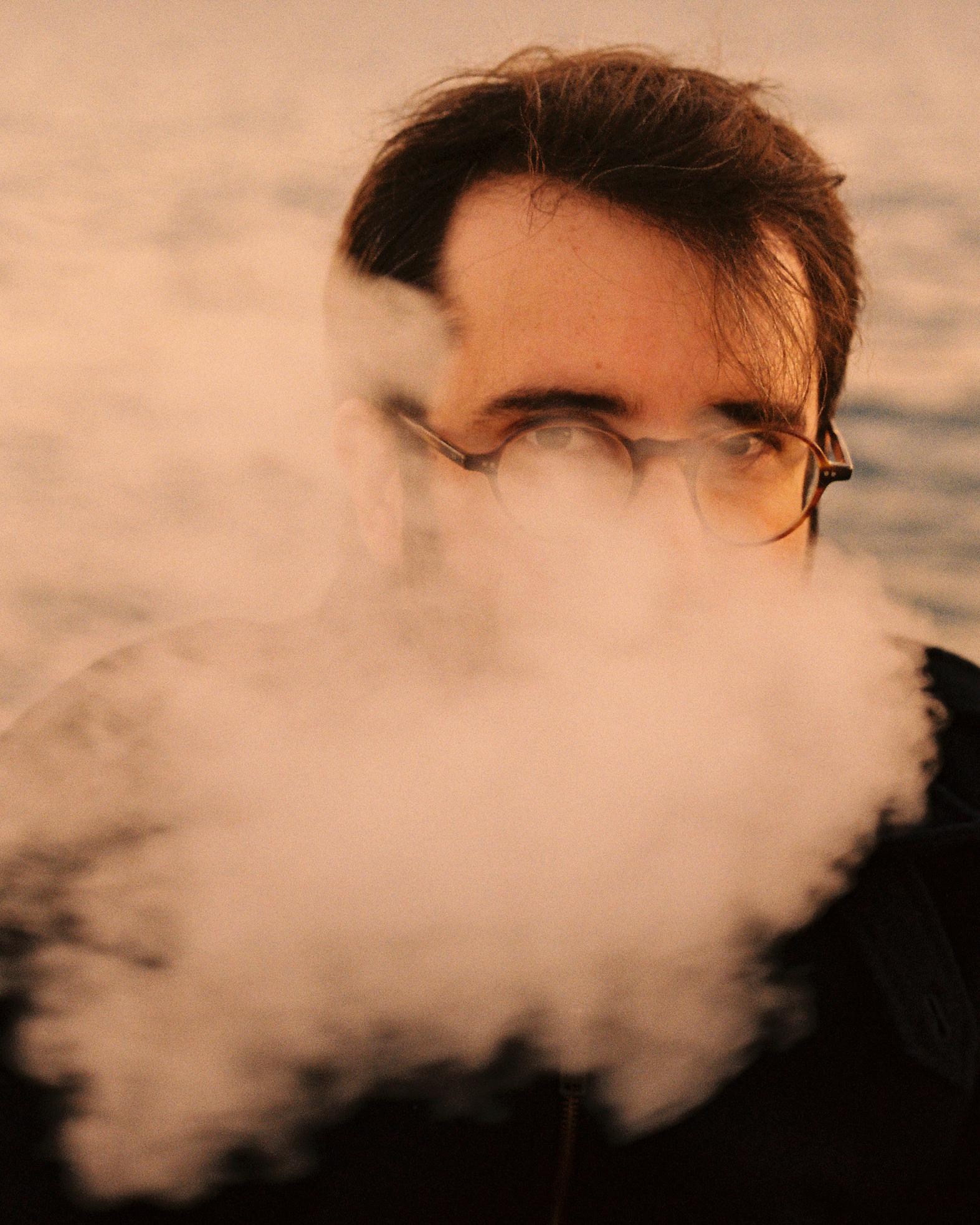 A man with a thick, white beard and a long, dark coat, standing in front of a blurred, snowy landscape.