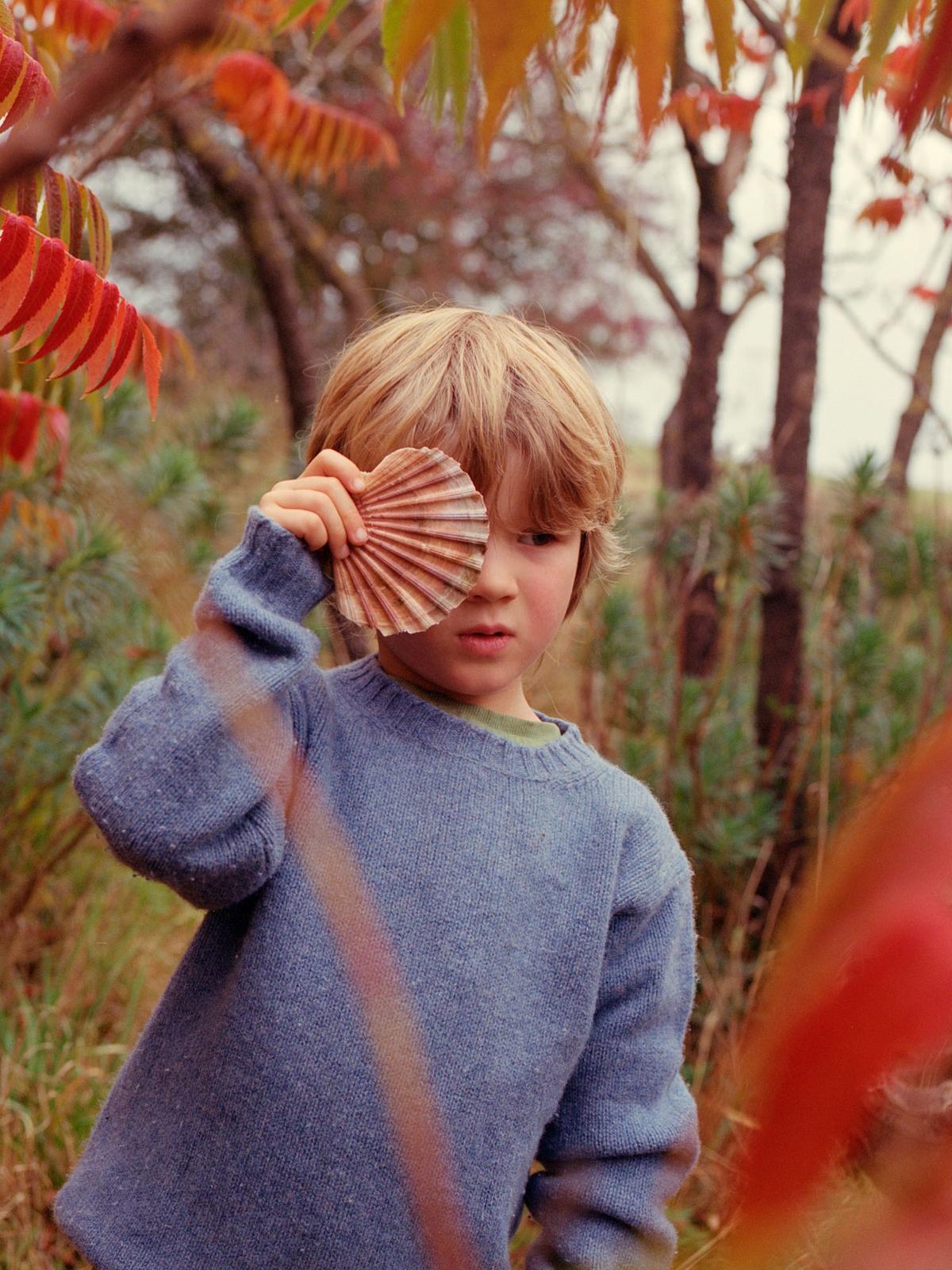 A young boy with blonde hair and a blue sweater holding a large, brown, fan-shaped shell up to his eye, in a natural, outdoor setting with trees and foliage in the background.