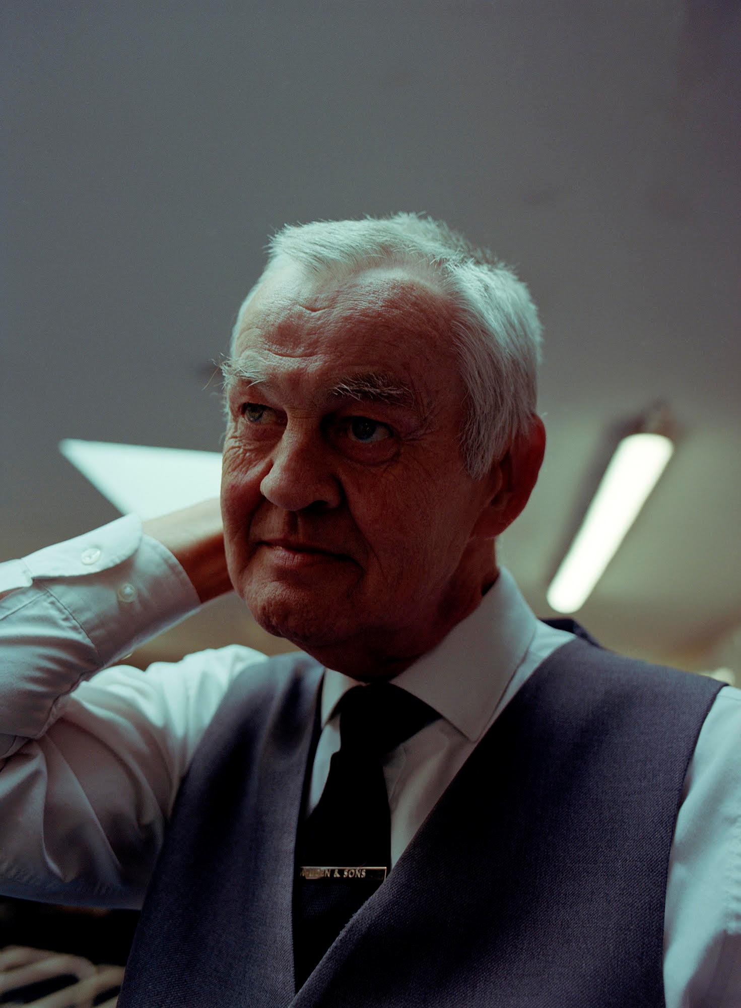 An older man with white hair, wearing a white shirt, black tie, and a grey vest, with his hand on his neck, in a dimly lit office setting.