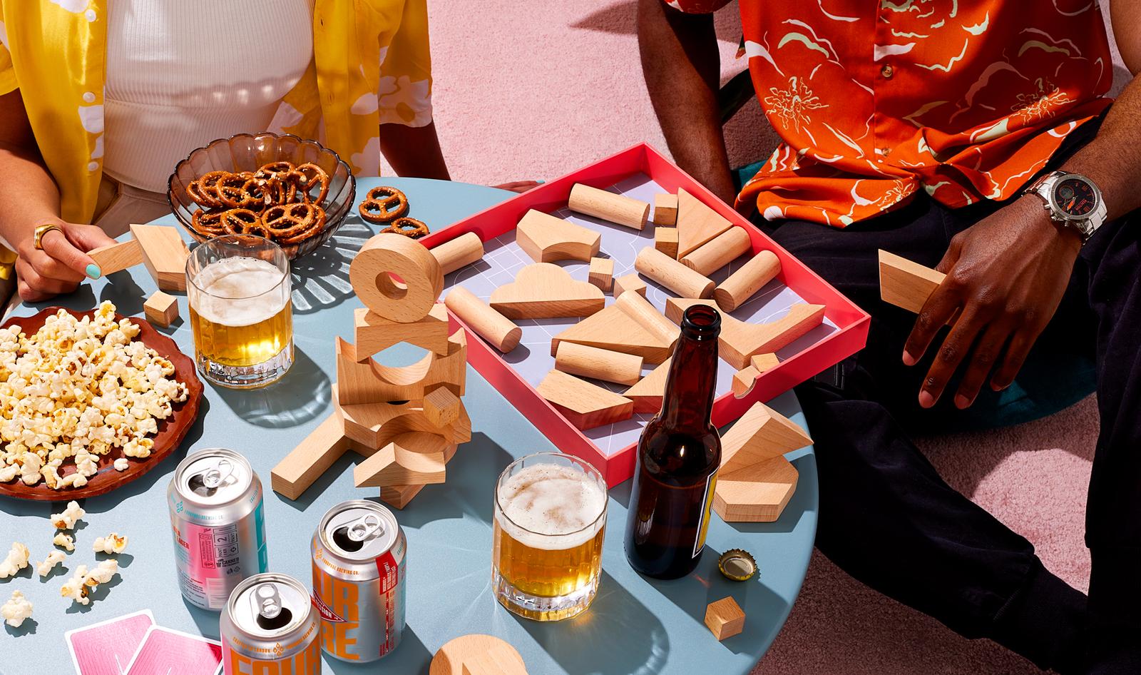 A woman and a man sit on the floor with a board game, snacks, and drinks surrounded by wooden blocks and cards.