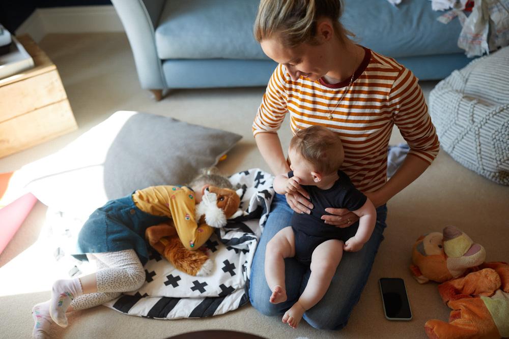 A woman sits on the floor with a baby in her lap, surrounded by toys and pillows. She looks down at the baby with a warm, loving expression.